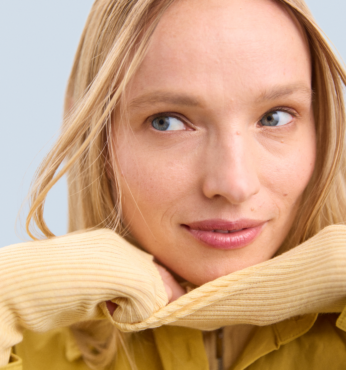 model with blonde hair looking to the left of the screen, wearing a mustard yellow sweater